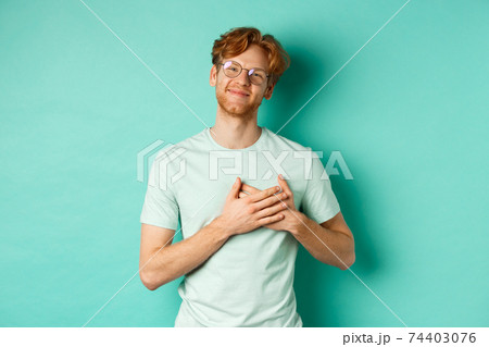 Handsome young man with red hair and glasses, holding hands on heart and smiling, saying thank you, feeling grateful and touched, standing over turquoise background Handsome young man with red hair and glasses, holding hands on heart and smiling, saying thank you, feeling grateful and touched, standing over turquoise background 74403076