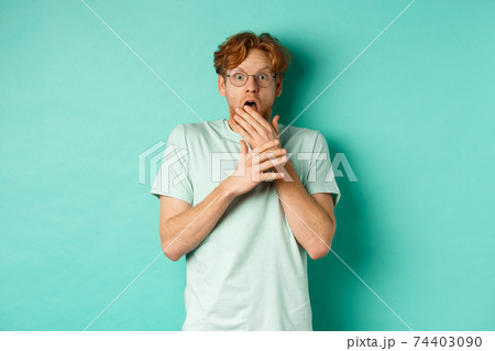 Shocked redhead man in glasses gasping startled, covering mouth and staring at camera scared, standing over turquoise background 74403090