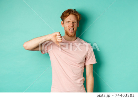 Disappointed redhead man showing thumbs-down and tongue, epxress dislike and disapproval, standing over turquoise background Disappointed redhead man showing thumbs-down and tongue, epxress dislike and disapproval, standing over turquoise background 74403583
