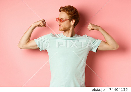 Confident young man with red hair, wearing summer sunglasses and t-shirt, showing strong and fit body muscles, flex biceps and staring cool at camera, pink background Confident young man with red hair, wearing summer sunglasses and t-shirt, showing strong and fit body muscles, flex biceps and staring cool at camera, pink background 74404136