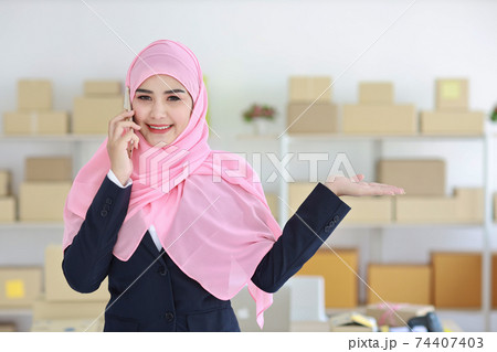 Religious asian muslim woman in blue suit and pink shaft on head standing and using mobile phone with confidence and showing something on hand with business package SME box delivery background. 74407403