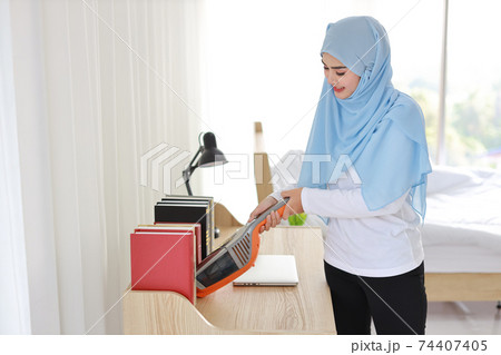 Active young asian muslim housewife woman cleaning with vacuum cleaning wooden table with computer blurred background. Cleaning girl in white dress and black leggings vacuuming her room 74407405