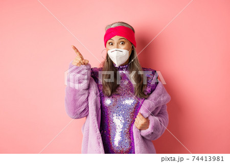 Covid and fashion concept. Amazed asian senior woman in glittering dress and face mask, pointing finger upper left corner, showing promo offer, pink background 74413981