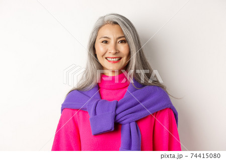 Close-up of beautiful asian senior woman in trendy pink sweater, smiling cheerful at camera, standing over white background Close-up of beautiful asian senior woman in trendy pink sweater, smiling cheerful at camera, standing over white background 74415080