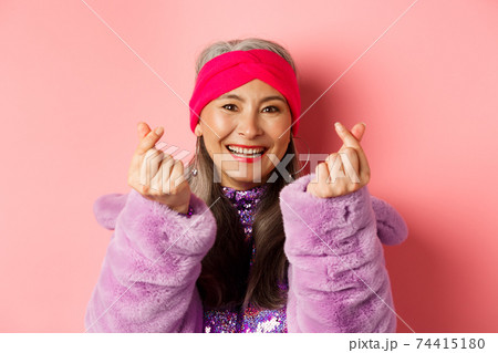 Fashion. Beautiful and fashionable asian senior woman smiling, showing korean heart gesture and looking happy, standing over pink background Fashion. Beautiful and fashionable asian senior woman smiling, showing korean heart gesture and looking happy, standing over pink background 74415180