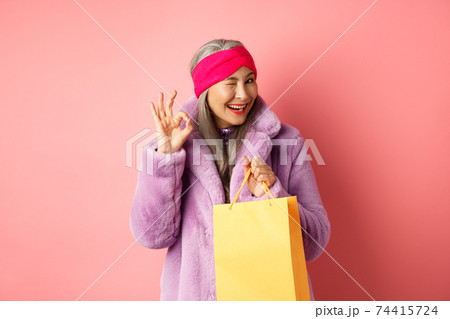 Stylish senior asian woman recommending store, holding shopping bag and winking with OK sign, smiling satisfied, standing over pink background Stylish senior asian woman recommending store, holding shopping bag and winking with OK sign, smiling satisfied, standing over pink background 74415724
