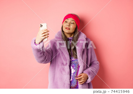 Fashion concept. Stylish asian senior female taking selfie on smartphone, posing in purple faux fur coat and party dress, standing over pink background Fashion concept. Stylish asian senior female taking selfie on smartphone, posing in purple faux fur coat and party dress, standing over pink background 74415946