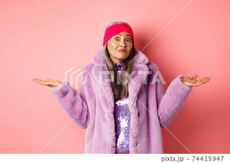 Fashionable and funky asian old woman shrugging shoulders, looking confused and clueless, standing in stylish purple coat and headband, pink background Fashionable and funky asian old woman shrugging shoulders, looking confused and clueless, standing in stylish purple coat and headband, pink background 74415947