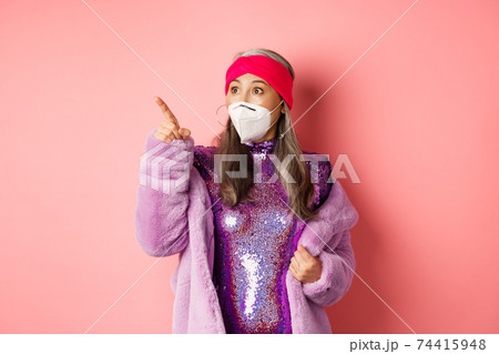 Covid and fashion concept. Impressed asian elderly woman checking out promo offer, wearing respirator from coronavirus, pointing and looking left, pink background 74415948