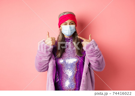 Covid-19, social distancing and fashion concept. Smiling asian senior woman in fashionable clothes and respirator, pointing at face mask and show thumb-up, pink background Covid-19, social distancing and fashion concept. Smiling asian senior woman in fashionable clothes and respirator, pointing at face mask and show thumb-up, pink background 74416022