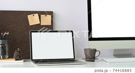 A white blank screen laptop is putting on a white working desk surrounded by office equipment. 74416883