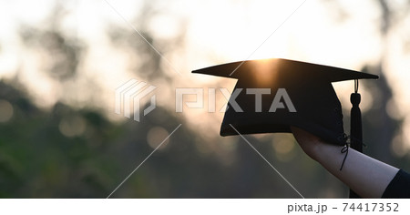 Cropped image of university student hand holding a graduation hat in hand over outdoors with sunset as background. Cropped image of university student hand holding a graduation hat in hand over outdoors with sunset as background. 74417352