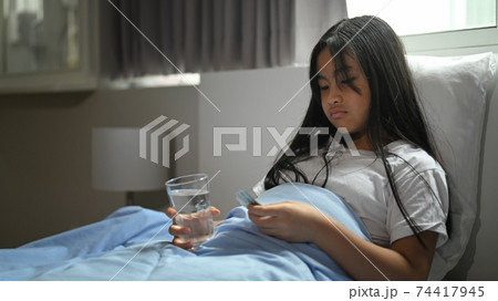 Sick little girl covered in a blanket is lying on the bed and taking a pill. 74417945