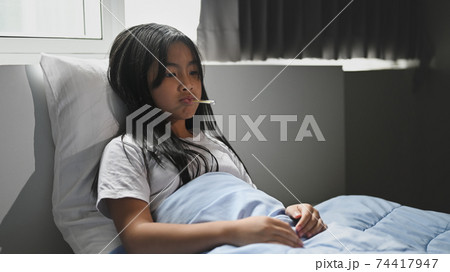 A little sick girl covered in a blanket is lying on the bed and measuring the temperature. A little sick girl covered in a blanket is lying on the bed and measuring the temperature. 74417947