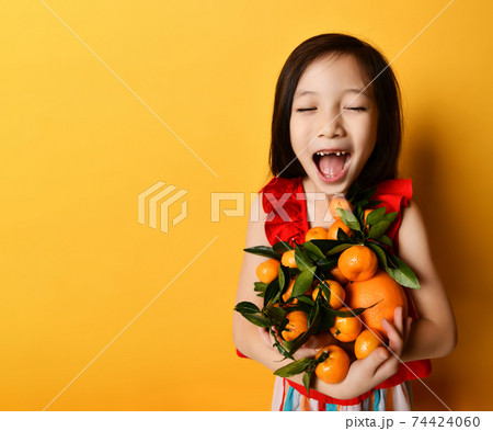 Asian child in red blouse. Looking wondered, holding an armful of tangerines and oranges, posing on orange background. Close up 74424060