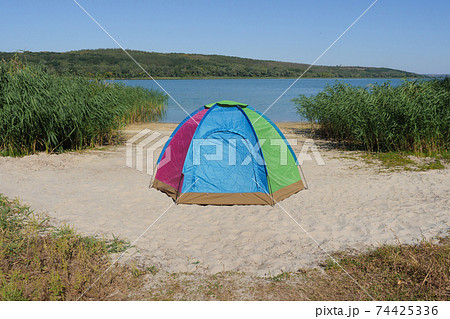 Tourist tent for trekking eco travel or fishing. Colourful dome tent on lake shore or camping equipment travel. Nobody near camping tent located on sand bank river in local hike travel solitude nature 74425336