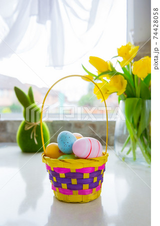 Straw bright basket with colored easter eggs, bouquet of yellow tulips and daffodils flowers and green bunny rabbit on white kitchen table near window. Festive spring vertical card. Selective focus. 74428058