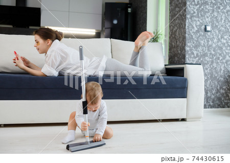 Baby boy play with mop, while mother surfing in your smartphone lying on couch Baby boy play with mop, while mother surfing in your smartphone lying on couch 74430615