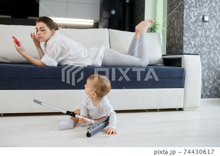 Baby boy play with mop, while mother surfing in your smartphone lying on couch Baby boy play with mop, while mother surfing in your smartphone lying on couch 74430617