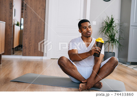African-American man sitting cross-legged and relaxing on exercise mat after workout, drinking water 74433860