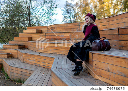 A smiling girl sits on a summer theater bench, made of wood in a burgundy coat and biret, an adult with a beautiful smile, in the fall against the background of trees, the blue sky. A smiling girl sits on a summer theater bench, made of wood in a burgundy coat and biret, an adult with a beautiful smile, in the fall against the background of trees, the blue sky. 74438076