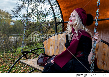A joyful lady sits on a garden swing, forged in a burgundy coat and biret, an adult looking at the camera, in the fall against the background of a pond, a blue sky. A joyful lady sits on a garden swing, forged in a burgundy coat and biret, an adult looking at the camera, in the fall against the background of a pond, a blue sky. 74438077
