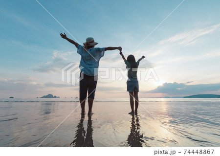 Silhouette of asian mother and daughter holding hand together standing on the beach looking at the beautiful sea and sky at the sunset time. Silhouette of asian mother and daughter holding hand together standing on the beach looking at the beautiful sea and sky at the sunset time. 74448867