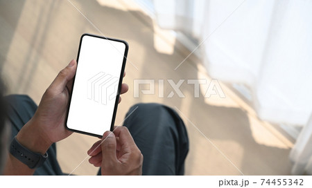 Close up view of young man holding mock up mobile phone with blank screen while sitting near window at home. 74455342