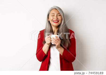 Business people concept. Happy office lady in red blazer enjoying drinking coffee, holding mug and smiling delighted, standing over white background Business people concept. Happy office lady in red blazer enjoying drinking coffee, holding mug and smiling delighted, standing over white background 74459189