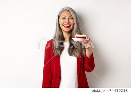 Celebration and holidays concept. Smiling asian businesswoman congratulate employees, giving plate with sweet cake and looking happy, standing over white background 74459258