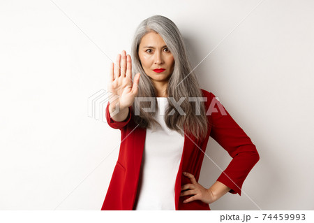 Serious asian woman in red blazer showing stop sign, telling no, frowning and rejecting something bad, standing against white background 74459993