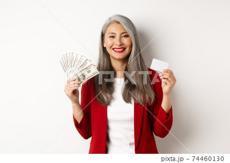 Successful asian senior businesswoman showing money in dollars and plastic card, smiling happy at camera, wearing red blazer and make-up 74460130