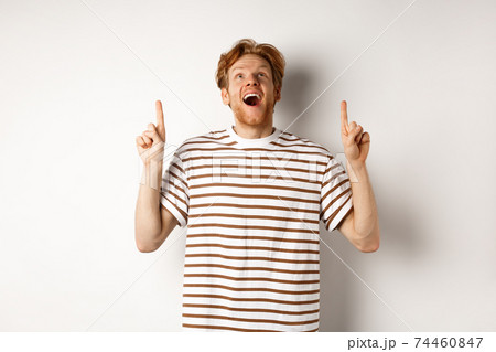 Fascinated redhead man looking with amazement and happiness, pointing fingers up, checking out something cool, standing over white background 74460847