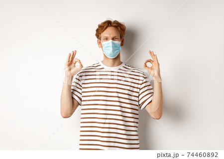 Covid-19, pandemic and social distancing concept. Young man with red hair wearing medical mask to prevent catching coronavirus, showing okay signs in approval, white background Covid-19, pandemic and social distancing concept. Young man with red hair wearing medical mask to prevent catching coronavirus, showing okay signs in approval, white background 74460892