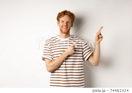 Handsome redhead man showing logo, pointing fingers right and smiling, standing over white background 74461324