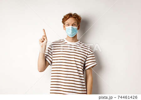 Covid, virus and social distancing concept. Surprised redhead man in face mask showing advertisement on top, pointing finger up and look amazed, white background Covid, virus and social distancing concept. Surprised redhead man in face mask showing advertisement on top, pointing finger up and look amazed, white background 74461426