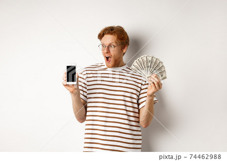 Amazed redhead man showing smartphone app on blank screen and money, winning prize cash online, standing over white background Amazed redhead man showing smartphone app on blank screen and money, winning prize cash online, standing over white background 74462988