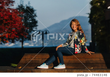 A happy little girl sits on a bench in a Sunny autumn sunset in the old town of Austria. A girl poses in a town against the backdrop of the Austrian Alps.Europe.Velden am w rthersee 74463146