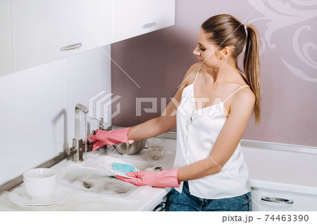housewife girl in pink gloves washes dishes by hand in the sink with detergent. The girl cleans the house and washes dishes in gloves at home. housewife girl in pink gloves washes dishes by hand in the sink with detergent. The girl cleans the house and washes dishes in gloves at home. 74463390