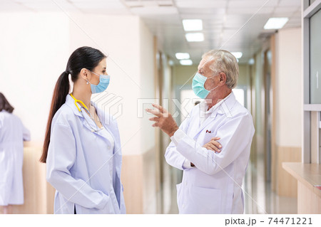 Caucasian senior male doctor wearing medical protective mask discussing of patient treatment case with female doctor at hospital corridor during Coronavirus epidemic 74471221
