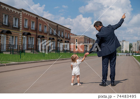 Rear view of a man in a black suit walks with his little daughter outdoors. A little girl in a white dress holds her father's hand. Paternal love 74483495