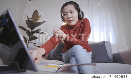 Asian woman on redshirt using a tablet with headphone for meeting online at home 74499653