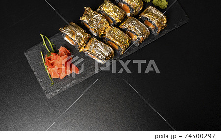Custom sushi roll with nori, cuttlefish ink, fresh salmon, cream cheese, pepper pumpkin, eel, unagi sauce and edible gold leaf with red ginger, wasabi on a square plate on black table and background. 74500297