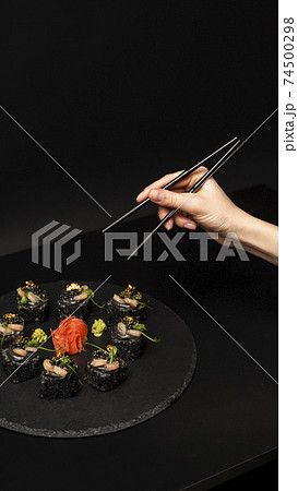 Hand with chopsticks wants to take custom sushi roll with black rice, crab meat, avocado, smoked salmon mousse, oar caviar, masago, shrimp cocktail, edible gold leaf, ginger, wasabi on black table. 74500298