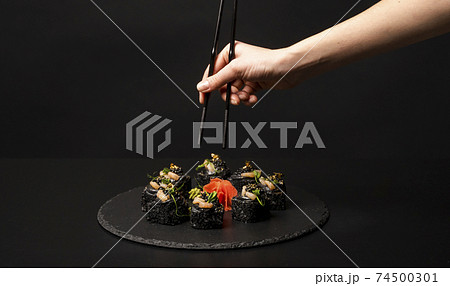 Hand with chopsticks wants to take custom sushi roll with black rice, crab meat, avocado, smoked salmon mousse, oar caviar, masago, shrimp cocktail, edible gold leaf, ginger, wasabi on black table. Hand with chopsticks wants to take custom sushi roll with black rice, crab meat, avocado, smoked salmon mousse, oar caviar, masago, shrimp cocktail, edible gold leaf, ginger, wasabi on black table. 74500301