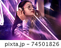 Neon Style. Asian girl in glossy jacket eyeglasses and headphones standing on the street in fluorescent violet lights listening to music looking up smiling joyful 74501826