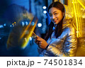 Neon Style. Asian girl in glossy jacket with wireless earphones standing on the street in fluorescent yellow lights listening to music browsing smartphone smiling cheerful 74501834