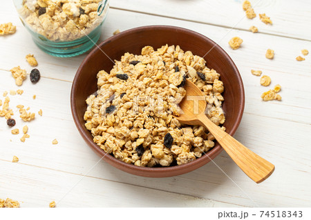 Muesli with dried grapes in a clay bowl on a white wooden table. Muesli is scattered on the table. Muesli with dried grapes in a clay bowl on a white wooden table. Muesli is scattered on the table. 74518343