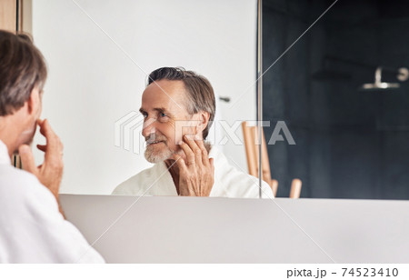 Smiling senior man looking at his reflection in the mirror and touching face. Mature male in a bathroom in the morning. Smiling senior man looking at his reflection in the mirror and touching face. Mature male in a bathroom in the morning. 74523410