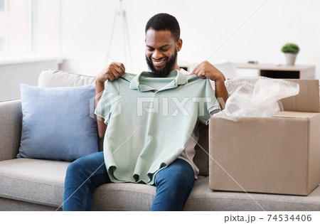 Happy Black Man Unpacking Box Holding New T-Shirt Sitting Indoors 74534406